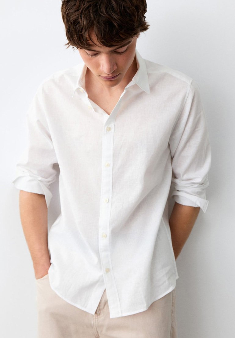 Jeune homme aux cheveux courts et bruns portant une chemise blanche boutonnée avec les manches retroussées et un pantalon beige clair, regardant vers le bas les mains dans les poches.