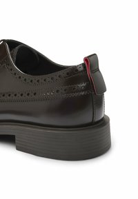 Scarpa nera in pelle con dettagli brogue, linguetta rossa a contrasto sul tallone e suola in gomma testurizzata. Presenta una forma arrotondata e una finitura liscia.