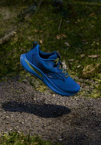 Mėlynas sportinis batelis su tekstūruotu tinklu, kontrastingomis geltonomis detalėmis ir paminkštintu padu, pakabintas virš natūralaus, žemės tonų fono.