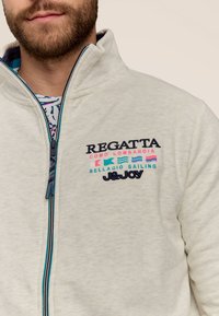 Lichtgrijze fleecetruit met rits, voorzien van marineblauwe en roze geborduurde logo's, hoge kraag en contrasterende blauwe ritsafwerking.