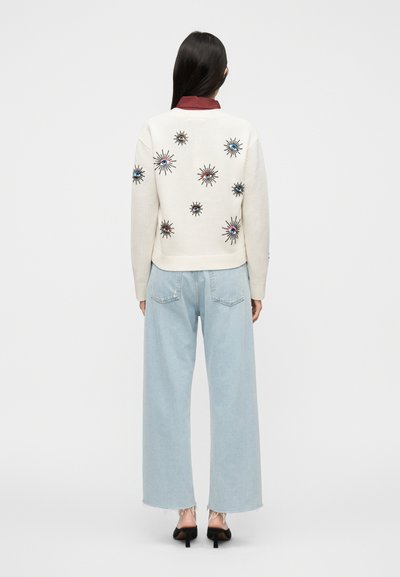 Πλεκτό εκρού πουλόβερ με πολύχρωμα κεντημένα μοτίβα εκρήξεων αστεριών, συνδυασμένο με ανοιχτόχρωμα τζιν φαρδιάς γραμμής και μαύρα mules με τακούνι.