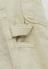 Béžová tkanina s dvojitě šitými švy, zobrazení detailu kapsy a tkaninového poutka na oděvu.