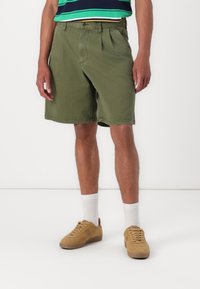 Olivegrüne Shorts aus Baumwolle mit einem lockeren Schnitt, ausgestattet mit vorderen Falten und seitlichen Taschen, kombiniert mit beigen Sneakers und weißen Socken.