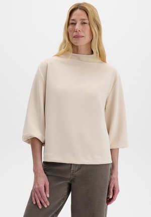 Donna con capelli biondi lunghi che indossa una maglia beige larga con maniche a tre quarti e pantaloni grigio scuro su sfondo uniforme.