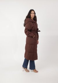 Abrigo largo marrón acolchado con cuello alto, cintura ceñida con cinturón y bolsillos laterales, combinado con vaqueros acampanados y sandalias de tacón transparente.