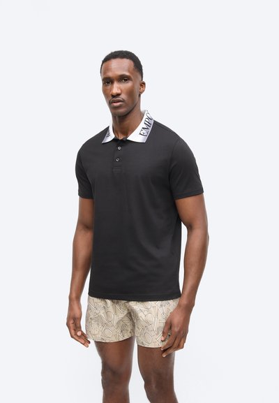 Άνδρας που φοράει μαύρο πόλο με λευκό γιακά και κείμενο, και μπεζ σορτς με μοτίβο από παντού, στέκεται μπροστά σε λευκό φόντο.