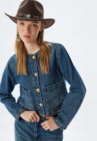 Chaqueta de mezclilla azul cortada con cierres de botones dorados, dos bolsillos frontales y mangas largas. Combinada con pantalones de mezclilla a juego y un sombrero marrón.