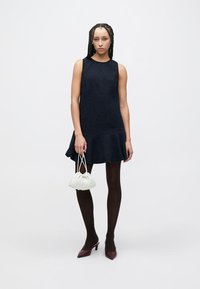 Mornarsko modra obleka brez rokavov s širokim robom in teksturirano tkanino, kombinirana s črnimi pajkicami in škornji bordo rdeče barve z zabrisanimi konicami. Majhna bela ročna torbica.