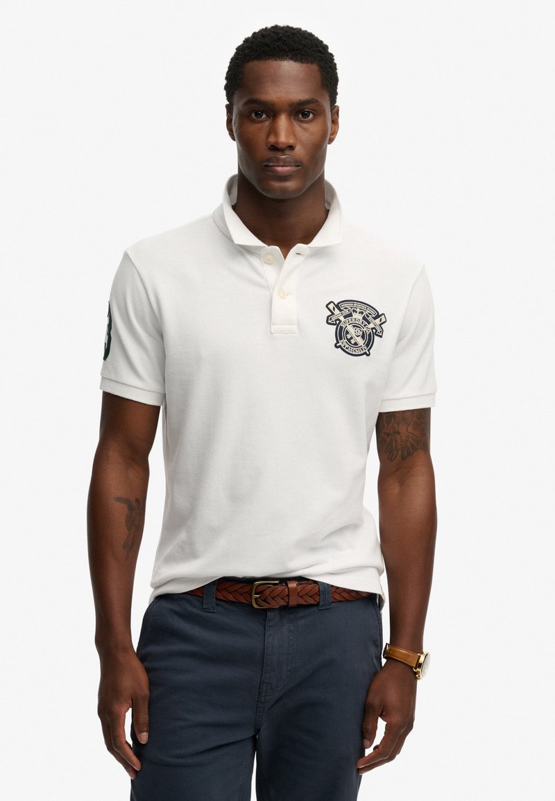 Weißes Poloshirt mit kurzen Ärmeln, gesticktem Logo auf der Brust, kombiniert mit dunkler Hose und einem braunen, geflochtenen Gürtel.