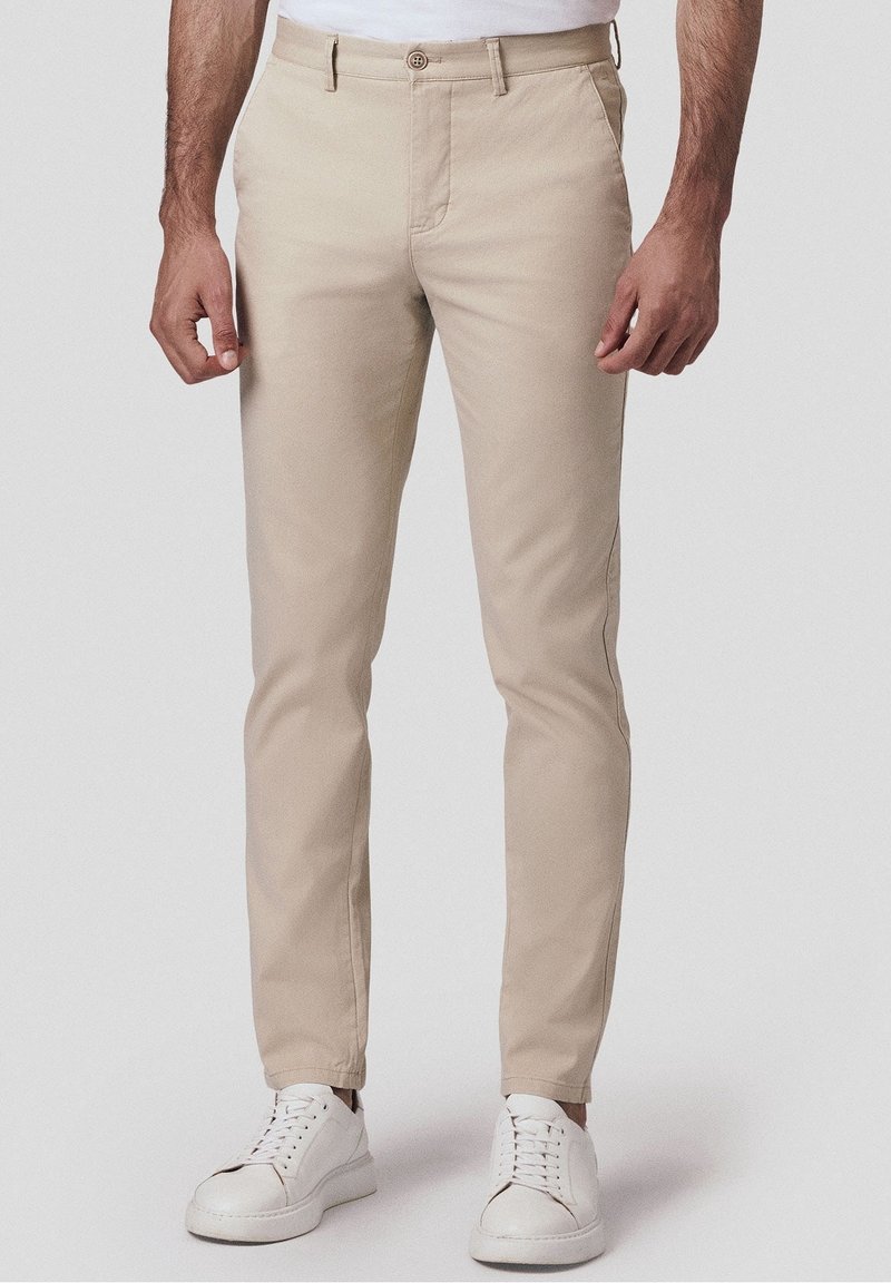 Homme portant un chino beige slim associé à des baskets blanches, debout devant un fond clair et uni.