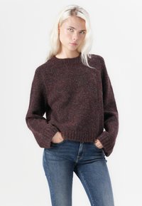 Maglione lavorato a maglia bordeaux con colletto rotondo, vestibilità rilassata e maniche ampie. Abbinato a jeans in denim blu per un look casual.