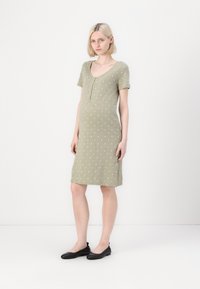 Olīvu zaļš, īsām piedurknēm grūtniecības kleita ar baltām punktiņām, ar apaļu izgriezumu un ceļa garuma malu. Apvienota ar melniem dzīvokļiem.