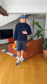 Νέος άνδρας με μπλε σκούρο μπουφάν, σκούρα σορτς, λευκές κάλτσες και αθλητικά παπούτσια στέκεται σε ένα σαλόνι κοντά σε ένα καφέ-κοκκινωπό καναπέ και ένα πράσινο φυτό.