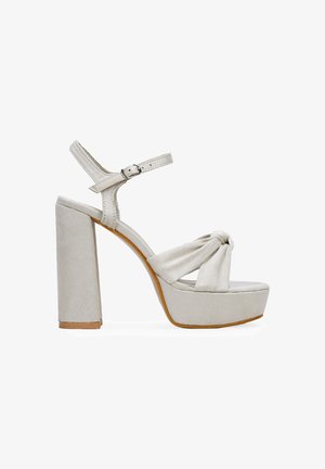 Beige platform sandal featuring a knotted suede upper, ankle strap with metal buckle, and a chunky high heel. Smooth texture and minimal design.