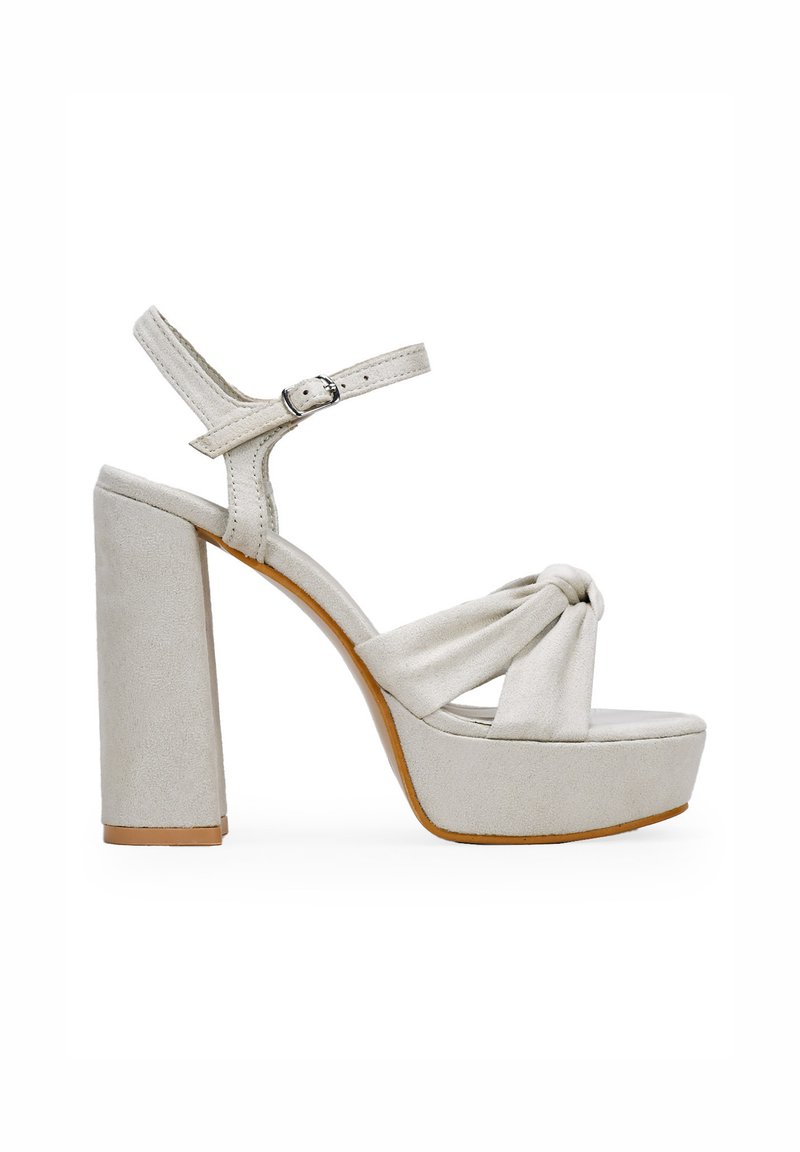 Beige platform sandal featuring a knotted suede upper, ankle strap with metal buckle, and a chunky high heel. Smooth texture and minimal design.