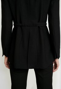 Blazer negro con cintura ajustada, diseño de espalda plisada, hombros estructurados y tela suave. Estilizado sobre pantalones negros ajustados.