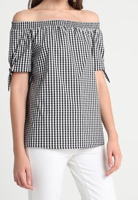 Black and white gingham off-the-shoulder blouse with short sleeves, gathered neckline, and tie accents, paired with white pants.