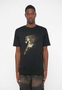Jeune homme portant un T-shirt noir avec un cheval en pixels dorés et le texte "Armani Exchange", accompagné de shorts cargo foncés.