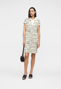 Γυναίκα που φοράει αμάνικο κρεμ φόρεμα με μαύρα αφηρημένα σχέδια, κρατώντας μια μαύρη στρογγυλή τσάντα και φοράει μαύρα ανοιχτά σανδάλια με τακούνι.