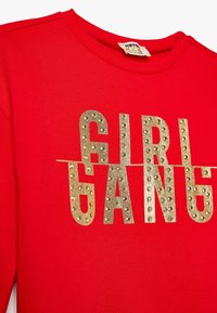 Felpa rossa in cotone con la scritta "GIRL GANG" in grandi lettere dorate testurizzate, arricchita da borchie metalliche. Design con scollatura a girocollo, morbida al tatto.