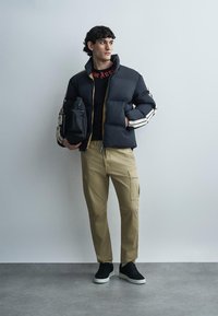 Junger Mann in schwarzer Steppjacke mit weißen Streifen, beigen Cargohosen, schwarzen Sneakern, hält eine schwarze Tasche, steht vor einer einfarbigen grauen Wand.