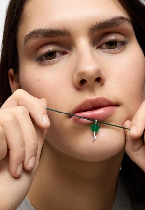 Joven sosteniendo un collar de cordón verde con un pequeño colgante de llave plateada cerca de sus labios, mostrando una expresión neutral.