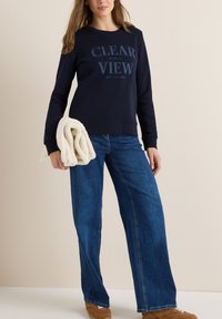 Sweat-shirt bleu marine avec le texte "CLEAR VIEW", associé à un jean large bleu clair. Le modèle tient une couverture texturée de couleur crème.