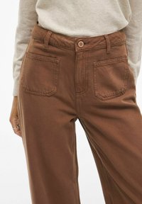 Pantaloni in cotone marrone con vita alta, passanti per cintura, chiusura frontale a bottone e due tasche posteriori. Presentano un tessuto liscio e strutturato.