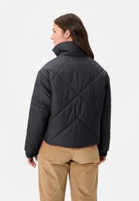 Veste matelassée noire avec col montant, motif losanges en piqué, et poignets élastiqués, associée à un pantalon en velours côtelé beige.