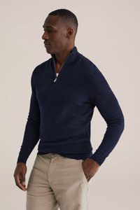 Tumši zils puszipa džemperis ar teksturētu adījumu, ribotu aproci un augstu apkakli, kopā ar gaiši bēšām biksēm.