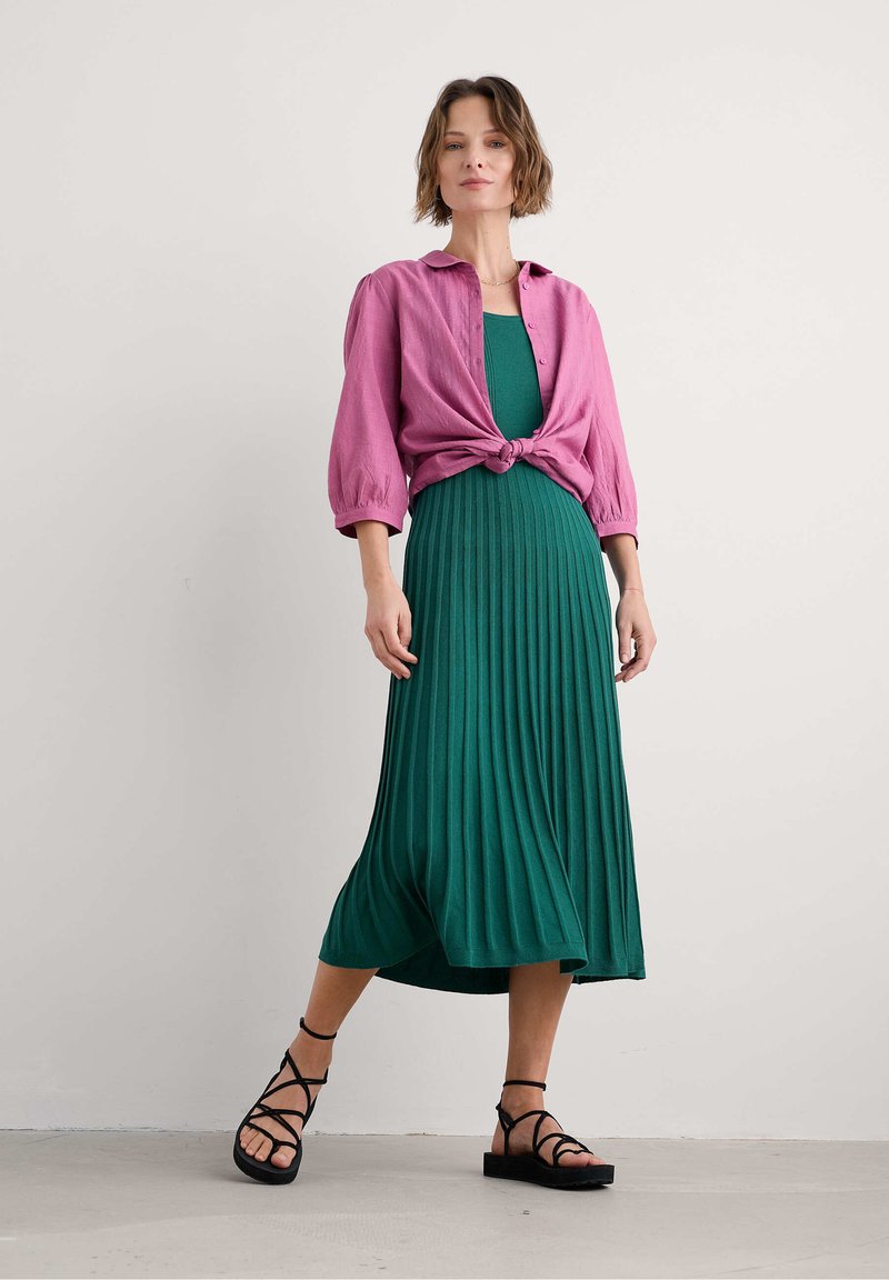 Green pleated midi dress paired with a knotted, buttoned light pink shirt. Black strappy sandals complete the outfit.