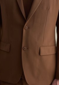 Giacca marrone con soffietto, due bottoni e tasche laterali, con una texture liscia e una vestibilità sartoriale.
