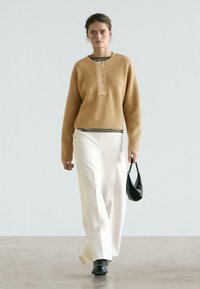 Pull marron côtelé avec patte de boutonnage, associé à un pantalon blanc taille haute à jambes larges. Tenant un petit sac à main noir, portant des bottes noires.