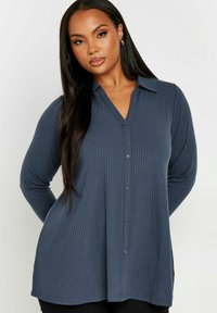 Mörkblå långärmad skjorta i ribbat tyg med V-ringning och kontrasterande knappar. Skjortan har en avslappnad passform och rundad nederkant.