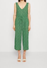 Groene bloemen jumpsuit met een V-hals, mouwloos ontwerp, verstelbare tailleband en cropped wijde pijpen. Draagt met crèmekleurige gevlochten sandalen.