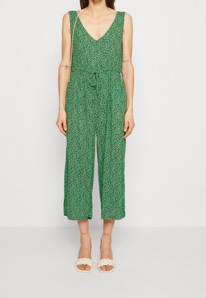 Combinaison florale verte avec un décolleté en V, sans manches, ceinture ajustable à la taille et jambes larges raccourcies. Portée avec des sandales crémeuses tressées.