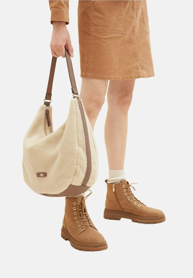 Beżowa torba na ramię z tkaniny o kremowej, puszystej teksturze, z skórzanymi akcentami i regulowanym paskiem. Brązowe botki na sznurówki z detalami zamka błyskawicznego i gumową podeszwą.