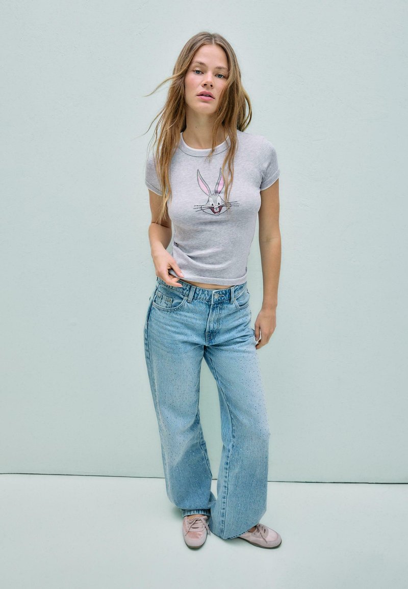 Mlada ženska z razpuščenimi dolgimi lasmi, oblečena v sivo majico z motivom Bugs Bunnyja, svetlo modre široke kavbojke in rožnate superge, stoji ob običajni steni.