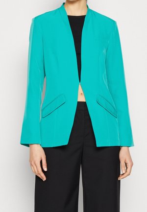 Person trägt einen taillierten türkisfarbenen Blazer über einem schwarzen Crop-Top und schwarzen Hosen, steht mit entspannten Armen seitlich.