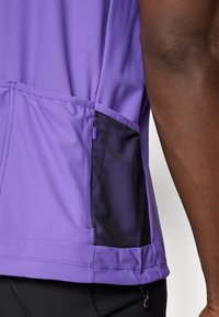 Camiseta deportiva morada con tejido texturizado, que cuenta con un bolsillo lateral de malla negra con cierre de cremallera y detalles de dobladillo elástico.