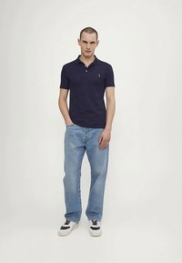 Άνδρας φορώντας ναυτικό πόλο μπλουζάκι, ανοιχτό γαλάζιο τζιν και λευκά αθλητικά παπούτσια, στέκεται με το ένα χέρι στην τσέπη, μπροστά σε ουδέτερο φόντο.