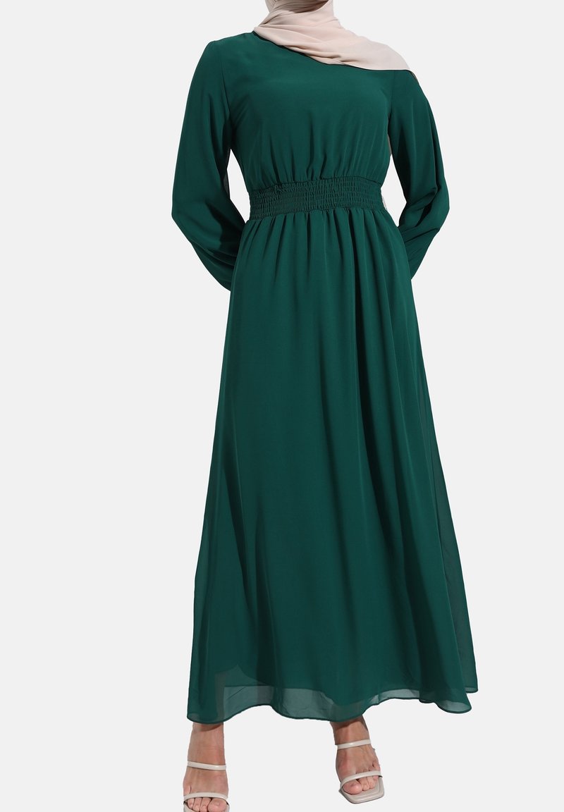 Femme portant une robe longue verte foncée à manches longues avec une taille froncée, associée à un foulard beige pâle et des sandales blanches ouvertes aux orteils.