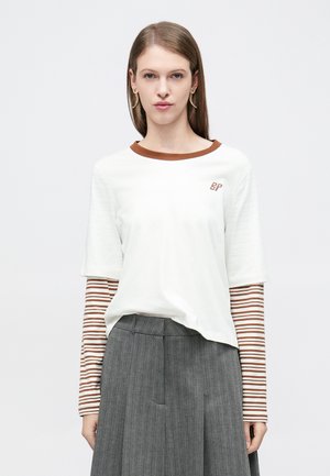 Biały t-shirt z krótkim rękawem i brązowym kołnierzem z logo "BP", noszony na paskowanym brązowo-kremowym longsleevie, zestawiony z szarą plisowaną spódnicą.