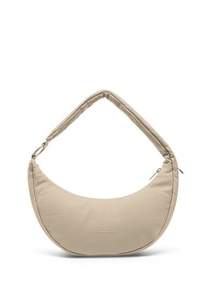 Jeune femme en t-shirt gris et pantalon noir tenant un sac à bandoulière beige en forme de croissant, posant devant un fond uni.