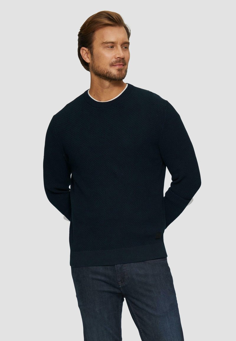 Pull en tricot bleu marine foncé avec un motif texturé, col rond, et poignets et ourlet côtelés. Porté sur un jean sombre.