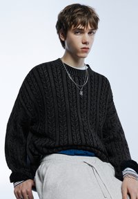 Hrubý čierny pletený sveter s dlhými rukávmi, nosený cez modré tričko, v kombinácii s svetlosivými teplákmi; doplnok strieborný retiazkový náhrdelník.
