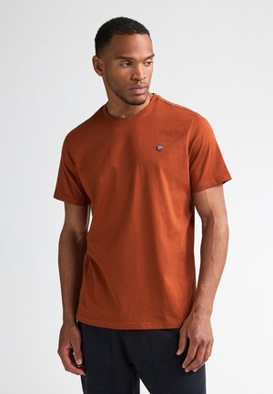 Man kijkt opzij, draagt een korte mouw T-shirt in verbrand oranje met een klein rond logo op de linkerborst en zwarte broek tegen een witte achtergrond.
