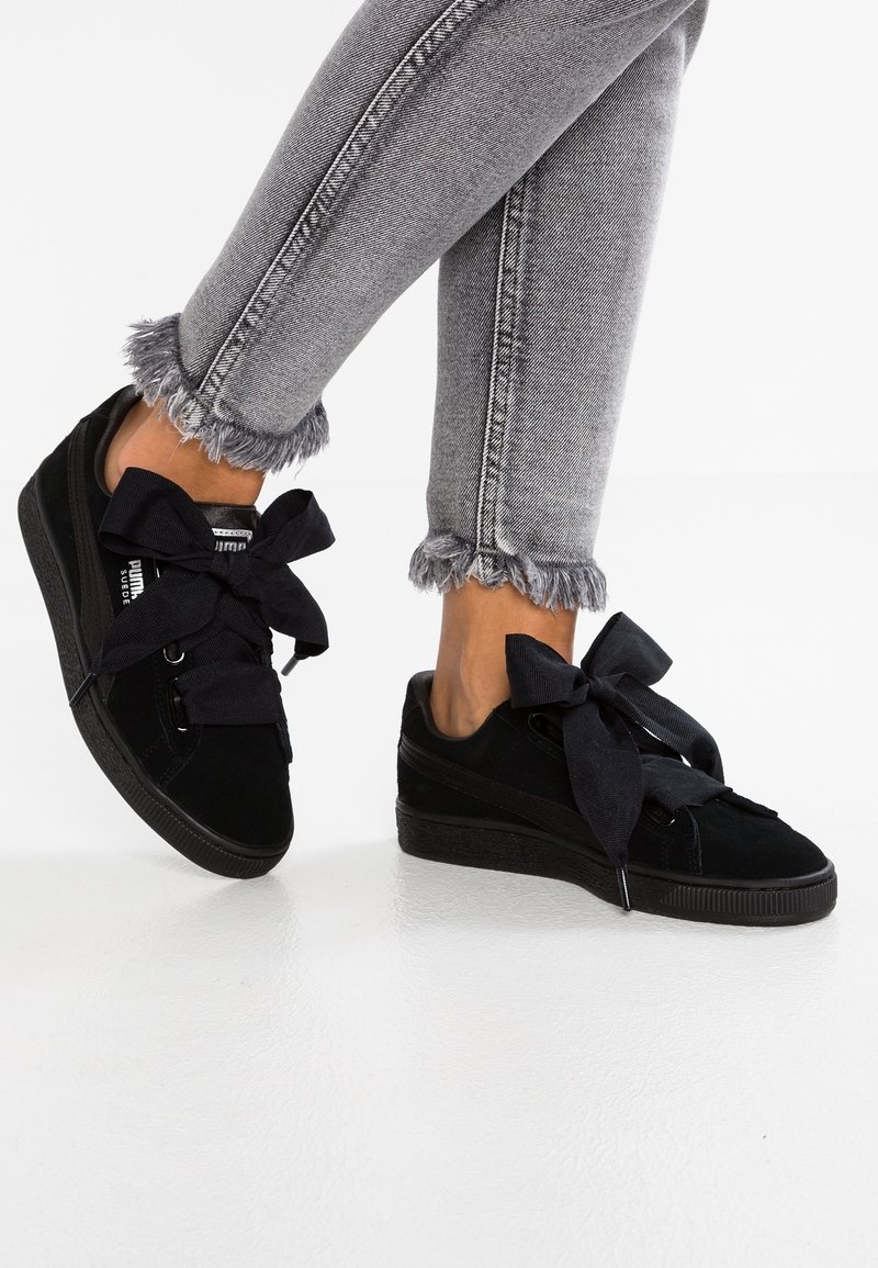 Jambes en jean gris à ourlet effiloché portant des baskets en daim noir Puma avec de grands lacets noirs en ruban sur un sol blanc.