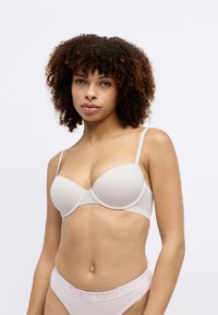 Jasno beżowy biustonosz z drutem, z formowanymi miseczkami i regulowanymi ramiączkami. Miękka tkanina z logo Calvin Klein na obwodzie, w zestawie z pasującymi majtkami.