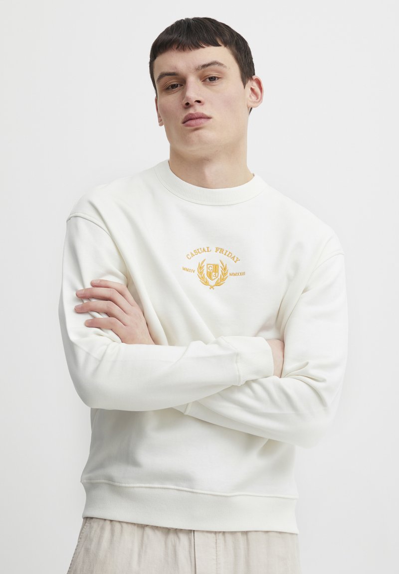 Sudadera blanca con cuello redondo acanalado, mangas con puños y un logo bordado "CASUAL FRIDAY" en dorado en el pecho. Textura suave.